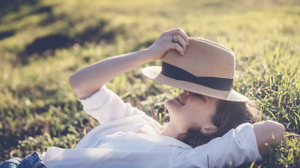 A woman in hat lying on green grass with closed eyes enjoying summer sun 