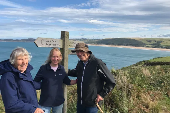 Three people standing by a coast path sign at Baggy Point looking across the sea on the North Devon coast