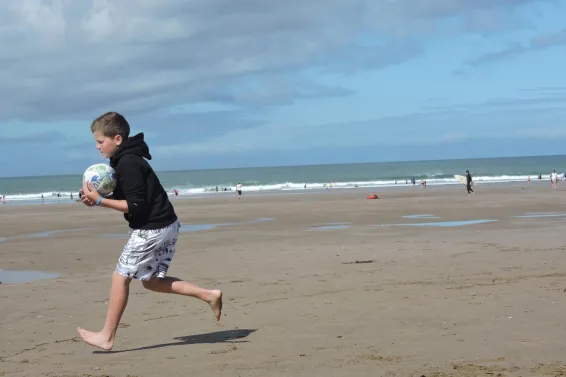 A boy running and holding a ball on the beach with the sea in the distance