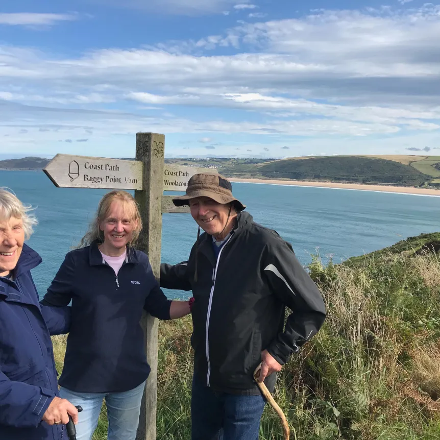 Three people standing by a coast path sign at Baggy Point looking across the sea on the North Devon coast