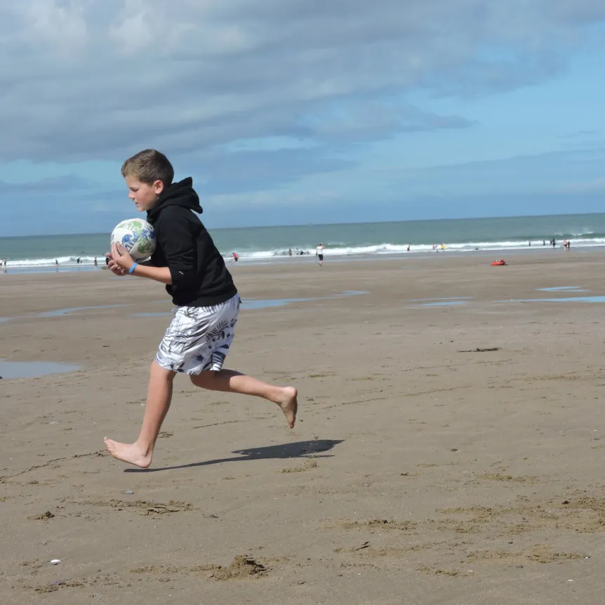 A boy running and holding a ball on the beach with the sea in the distance
