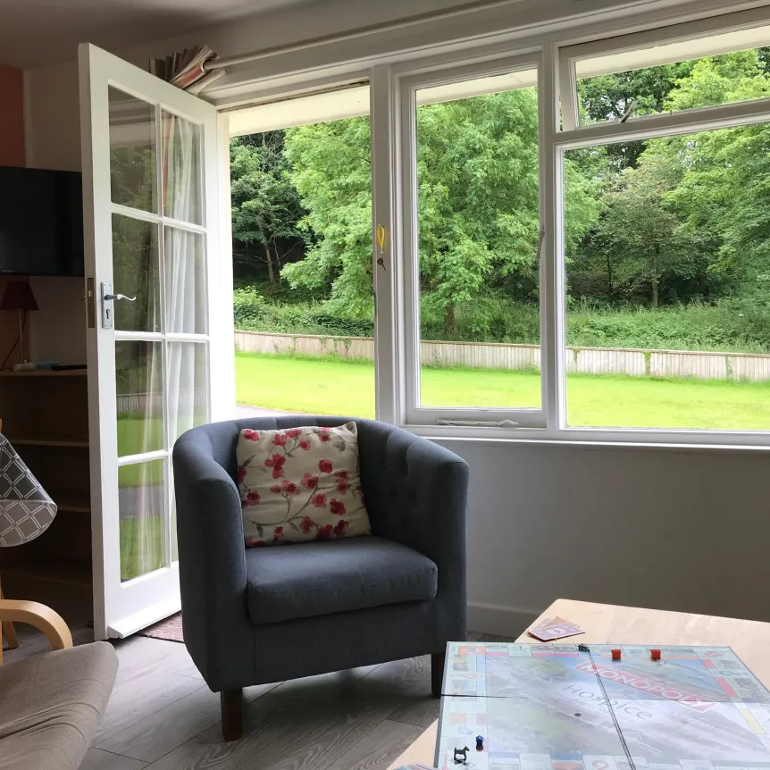 A chair in the doorway of the lounge inside Hibiscus Cottage with a view over the lawn at Spreacombe Holiday Cottages